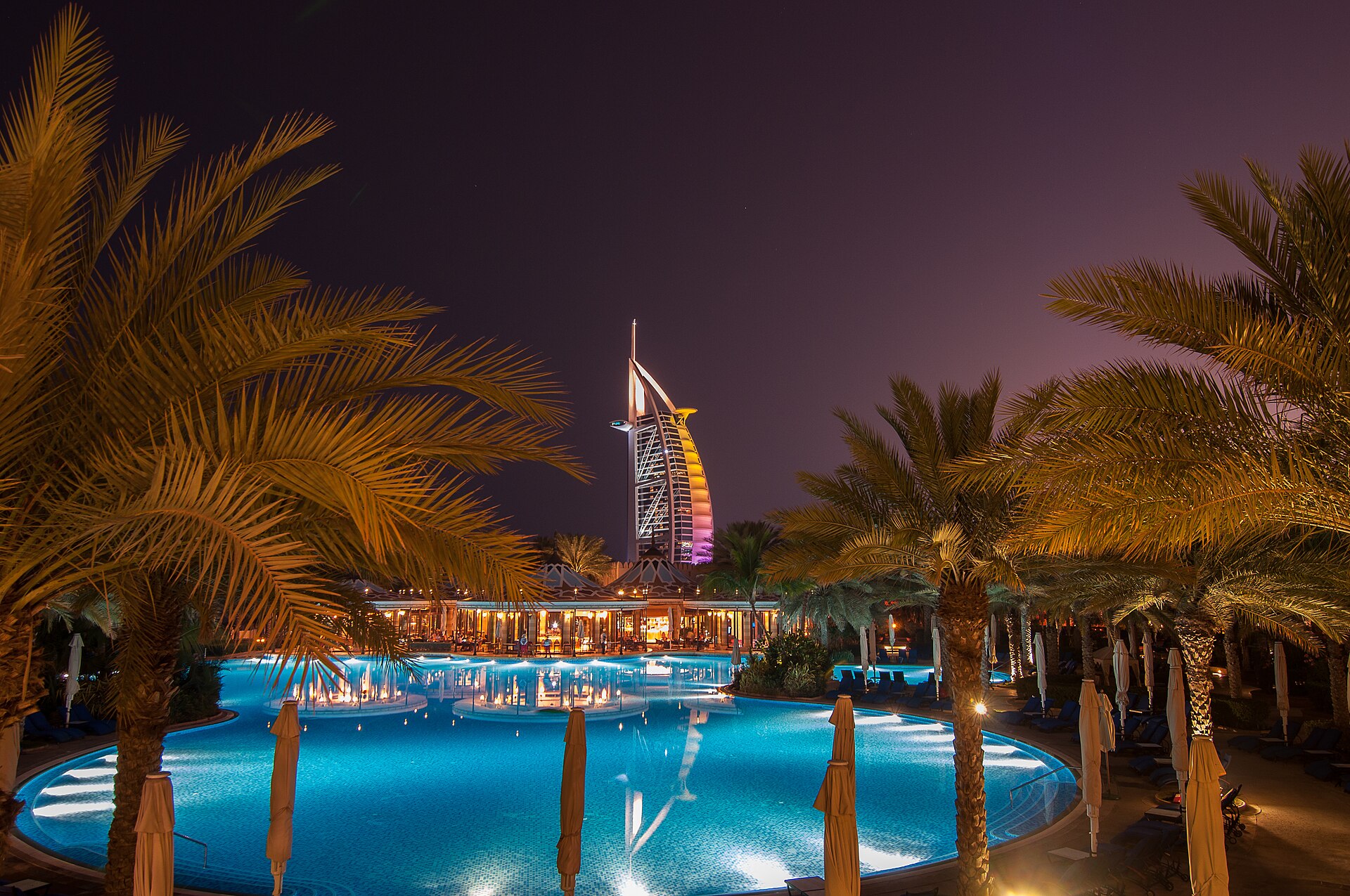 Burj Al Arab iluminado à noite visto entre palmeiras e piscina em Jumeirah